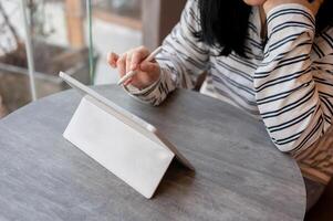 een Aziatisch vrouw gebruik makend van haar digitaal tablet, Holding een tablet pen, terwijl werken van een afstand Bij een cafe. foto