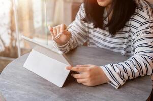 een Aziatisch vrouw gebruik makend van haar digitaal tablet, Holding een tablet pen, terwijl werken van een afstand Bij een cafe. foto