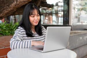 een glimlachen en positief Aziatisch vrouw is werken van een afstand buitenshuis in een stad park, gebruik makend van haar laptop. foto