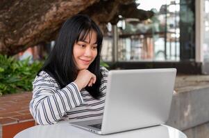 een glimlachen en positief Aziatisch vrouw is werken van een afstand buitenshuis in een stad park, gebruik makend van haar laptop. foto