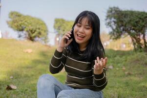 een vrolijk jong Aziatisch vrouw genieten van pratend Aan de telefoon met haar vriend terwijl ontspannende in een park. foto
