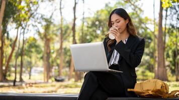 een elegant Aziatisch zakenvrouw werken van een afstand Bij een park, nippen koffie en werken Aan haar laptop. foto