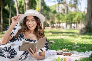 aantrekkelijk vrouw in een schattig jurk en een rietje hoed is lezing een boek Aan een picknick mat in een groen park. foto