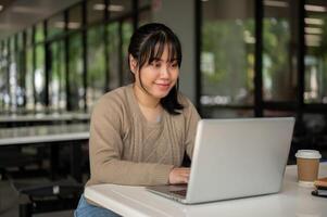 een jong Aziatisch vrouw college leerling werken van een afstand Bij een kantine, werken Aan haar laptop computer. foto