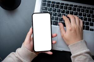 een top visie beeld van een vrouw typen Aan laptop toetsenbord en Holding een wit scherm smartphone model. foto