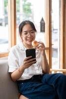 een jong, gelukkig Aziatisch vrouw genieten van een donut terwijl gebruik makend van haar smartphone Aan een sofa in een cafe. foto