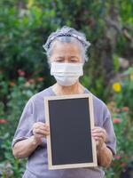 portret van ouderen vrouw staand Holding een zwart etiket en vervelend gezicht masker voor gezond omdat hebben lucht verontreiniging p.m 2.5. masker voor beschermen virus, bacteriën, stuifmeel granen. gezondheidszorg concept foto