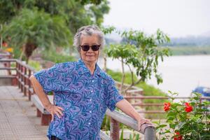 ouderen vrouw met kort wit haar- staand glimlachen en op zoek Bij de camera Bij kant de rivier. Aziatisch senior vrouw gezond en hebben positief gedachten Aan leven maken haar gelukkig elke dag. Gezondheid concept foto