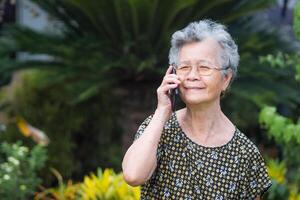 een portret van een senior vrouw gebruik makend van een smartphone en op zoek weg terwijl staand in een tuin. ruimte voor tekst. concept van oud mensen en technologie foto