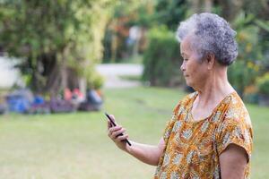 senior vrouw met kort grijs haar- Holding en op zoek Bij een smartphone terwijl staand in een tuin. ruimte voor tekst. concept van oud mensen en communicatie foto