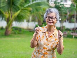 een gelukkig senior vrouw tonen balanceren vuisten opgewonden voor succes met armen verheven vieren zege glimlachen terwijl staand in de tuin. concept van oud mensen en gezondheidszorg foto