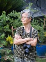 portret van een ouderen vrouw met kort grijs haar- armen gekruist, hangende een digitaal camera Aan de nek en op zoek uit naar de kant terwijl staand in een tuin. concept van oud mensen en fotografie foto