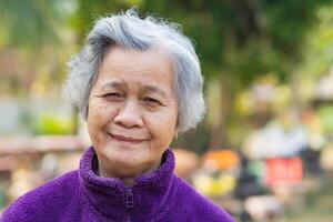 vrolijk ouderen Aziatisch vrouw met kort grijs haar, glimlachen en op zoek Bij de camera terwijl staand in een tuin. ruimte voor tekst. concept van oud mensen en gezondheidszorg foto