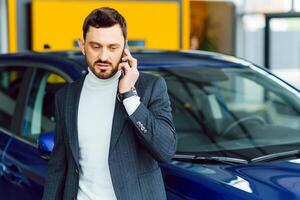 de auto shows de karakter van een Mens. zelfverzekerd succes elegant bedrijf Mens in vol pak en met telefoon in hand- staand in de buurt nieuw blauw auto looks weg foto