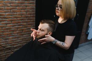 jong volwassen mooi Kaukasisch vrouw kapper bezuinigingen baard knap Mens Bij modern kapperszaak gelukkig homo zittend stoel mannen schoonheid salon. baard zorg. metroseksueel. vrouw kapper in zwart handschoenen portie. foto