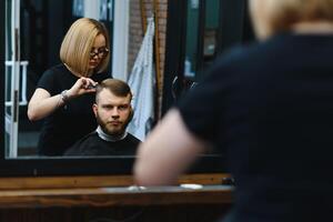 elegant Mens zittend kapper winkel haar stylist kapper vrouw snijdend zijn haar- portret knap gelukkig jong gebaard Kaukasisch vent krijgen modieus kapsel aantrekkelijk kapper meisje werken portie cliënt. foto