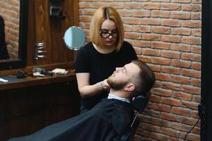 geschoold vrouw kapper snijdend een baard van brunette volwassen vent in professioneel retro kapperszaak. foto
