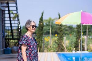 een portret van een ouderen vrouw vervelend zonnebril en rietje hoed terwijl staand kant zwemmen zwembad. ruimte voor tekst. concept van oud mensen en vakantie foto