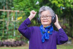 senior vrouw met kort grijs haar- vervelend bril en draadloze hoofdtelefoons luisteren naar een liedje, glimlachen terwijl staand in een tuin. concept van oud mensen en ontspanning foto