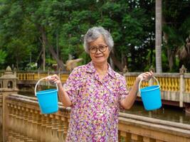 portret van een ouderen Aziatisch vrouw Holding een blauw plastic emmer, glimlachen en op zoek Bij de camera terwijl staand kant de vijver. concept van oud mensen en gezondheidszorg foto