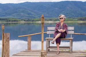 portret van senior Aziatisch vrouw vervelend zonnebril en op zoek Bij de camera terwijl zittend Aan een houten stoel kant de vijver. concept van oud mensen en ontspanning foto