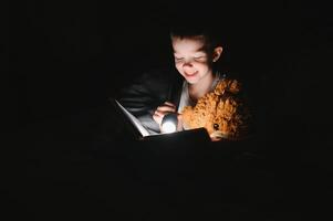 kind lezing boek in bed. kinderen lezen Bij nacht. weinig jongen met fee verhaal boeken in slaapkamer . onderwijs voor jong kinderen. bedtijd verhaal in de avond. schattig kind onder deken in donker kamer met lamp foto