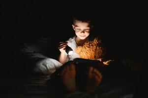 kind lezing boek in bed. kinderen lezen Bij nacht. weinig jongen met fee verhaal boeken in slaapkamer . onderwijs voor jong kinderen. bedtijd verhaal in de avond. schattig kind onder deken in donker kamer met lamp foto