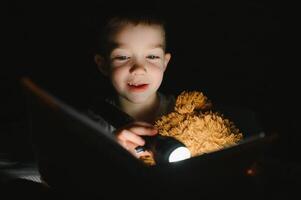 kind lezing boek in bed. kinderen lezen Bij nacht. weinig jongen met fee verhaal boeken in slaapkamer . onderwijs voor jong kinderen. bedtijd verhaal in de avond. schattig kind onder deken in donker kamer met lamp foto