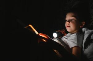 portret van schattig weinig jongen lezing in bed met zaklamp in donker kamer, genieten van sprookjes. foto