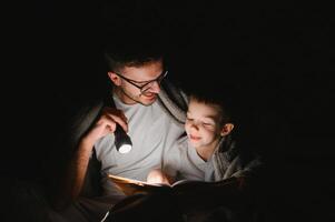 vader en zoon met zaklamp lezing boek onder deken Bij huis. foto