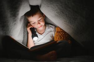 kind lezing boek in bed. kinderen lezen Bij nacht. weinig jongen met fee verhaal boeken in slaapkamer . onderwijs voor jong kinderen. bedtijd verhaal in de avond. schattig kind onder deken in donker kamer met lamp foto