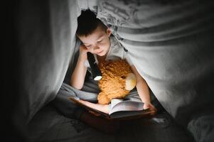 lezing boek en gebruik makend van zaklamp. jong jongen in gewoontjes kleren aan het liegen naar beneden in de buurt tent Bij avond tijd foto