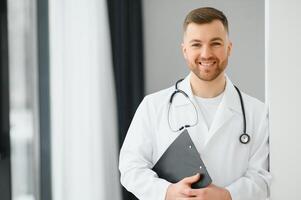 portret van knap jong dokter Aan ziekenhuis gang op zoek Bij camera, lachend. foto