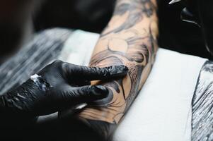 bijgesneden dichtbij omhoog van een gebaard tatoeëren artiest werken Bij zijn studio tatoeëren mouw Aan de arm van zijn mannetje cliënt. Mens krijgen getatoeëerd door professioneel tatoeëerder foto