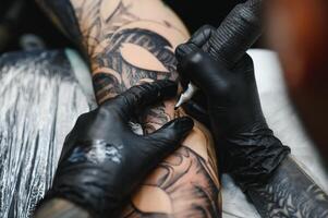 bijgesneden dichtbij omhoog van een gebaard tatoeëren artiest werken Bij zijn studio tatoeëren mouw Aan de arm van zijn mannetje cliënt. Mens krijgen getatoeëerd door professioneel tatoeëerder foto