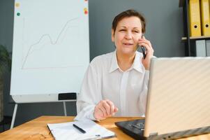 ouderen vrouw werken Aan laptop computer, pratend Aan de telefoon. foto