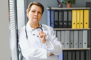 portret van midden- leeftijd vrouw dokter is vervelend een wit dokter jas met een stethoscoop in de omgeving van haar nek. foto