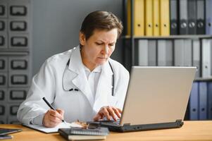 portret schot van midden- oud vrouw dokter zittend Bij bureau en werken in kantoor. foto