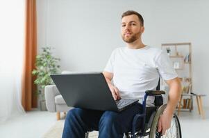 portret van glimlachen gehandicapt mannetje zittend in rolstoel en werken Aan laptop van huis. jong arbeider met speciaal behoeften. freelancer en mensen met handicaps concept. foto