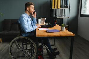 gehandicapt persoon in de rolstoel werken in de kantoor Bij de computer. hij is glimlachen en gepassioneerd over de workflow foto