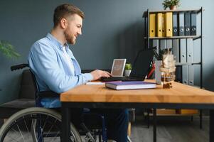 jong Mens met speciaal behoeften in gewoontjes kleren werken Aan draadloze laptop. mannetje freelancer werken van huis terwijl zittend in rolstoel. foto