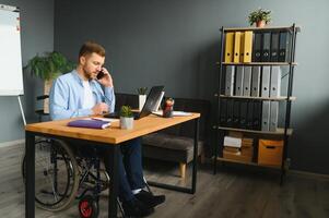 gehandicapt persoon in de rolstoel werken in de kantoor Bij de computer. hij is glimlachen en gepassioneerd over de workflow foto