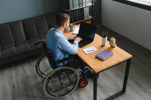 jong Mens met speciaal behoeften in gewoontjes kleren werken Aan draadloze laptop. mannetje freelancer werken van huis terwijl zittend in rolstoel. foto