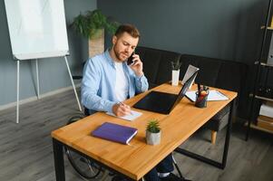 jong Mens met speciaal behoeften in gewoontjes kleren werken Aan draadloze laptop. mannetje freelancer werken van huis terwijl zittend in rolstoel foto