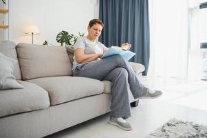 middelbare leeftijd huisvrouw zittend Aan sofa met boek, vrij tijd pensioen, hobby. foto