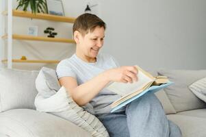 portret van een gelukkig volwassen vrouw ontspannende Aan bankstel en lezing een boek in een modern huis foto