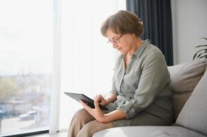 senior vrouw gebruik makend van digitaal tablet in leven kamer Bij huis foto