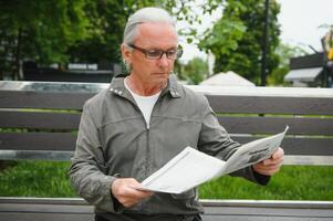 portret van senior Mens lezing Aan bank gedurende zomer dag. foto