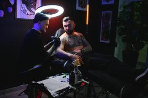 gebaard tatoeëren artiest werken Bij zijn studio tatoeëren mouw Aan de arm van zijn mannetje cliënt. Mens krijgen getatoeëerd door professioneel tatoeëerder foto