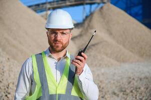 een arbeider in een moeilijk hoed staat met een walkietalkie Bij een steen verpletterend fabriek. foto